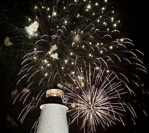Fireworks burst over a lighthouse against a dark night sky.
