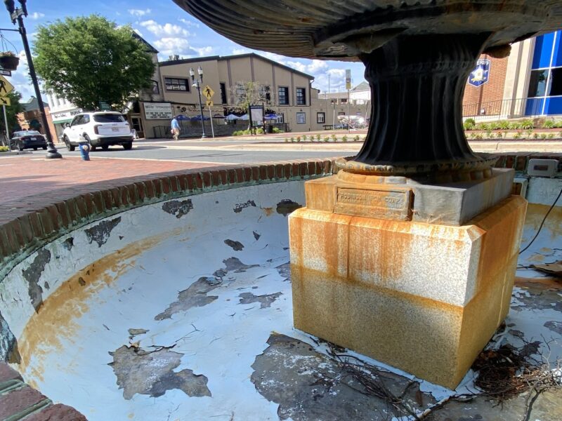 Empty, peeling fountain basin with a central pedestal in a sunny downtown square.