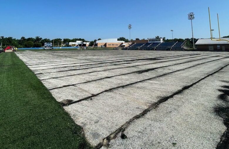 Empty sports field covered with white tarp beside stadium bleachers under a blue sky.