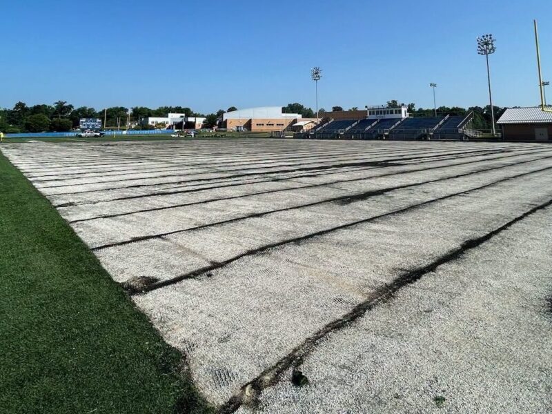 Empty sports field covered with white tarp beside stadium bleachers under a blue sky.