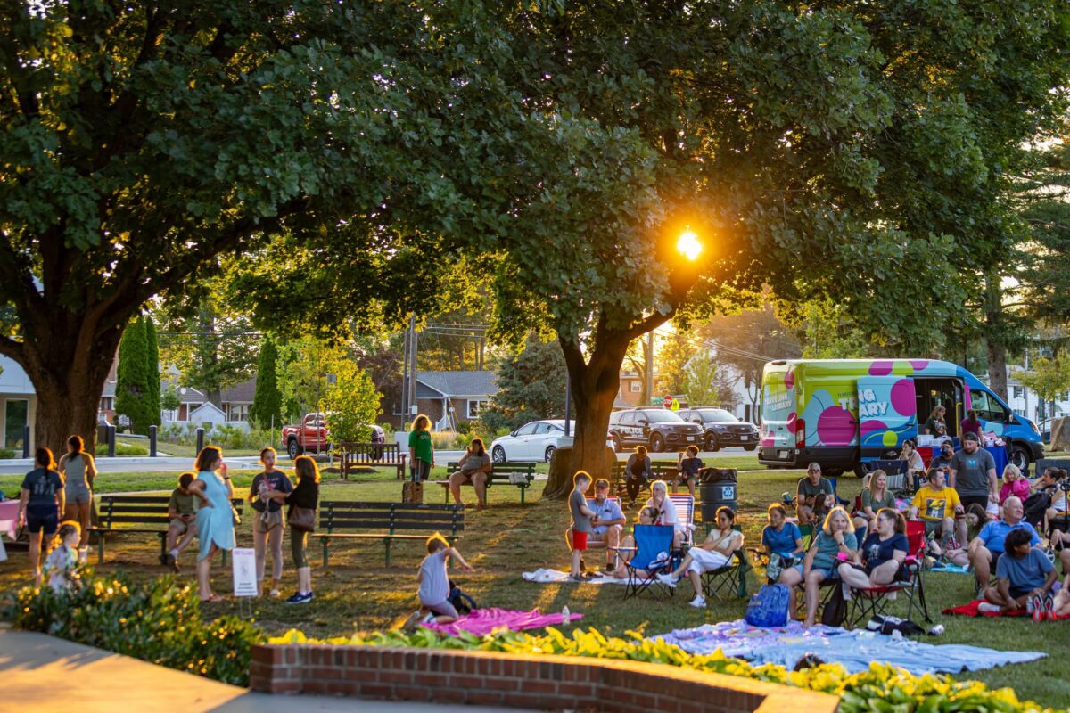 Crowd gathers on a grassy park lawn at sunset beside a colorful van and picnic blankets.