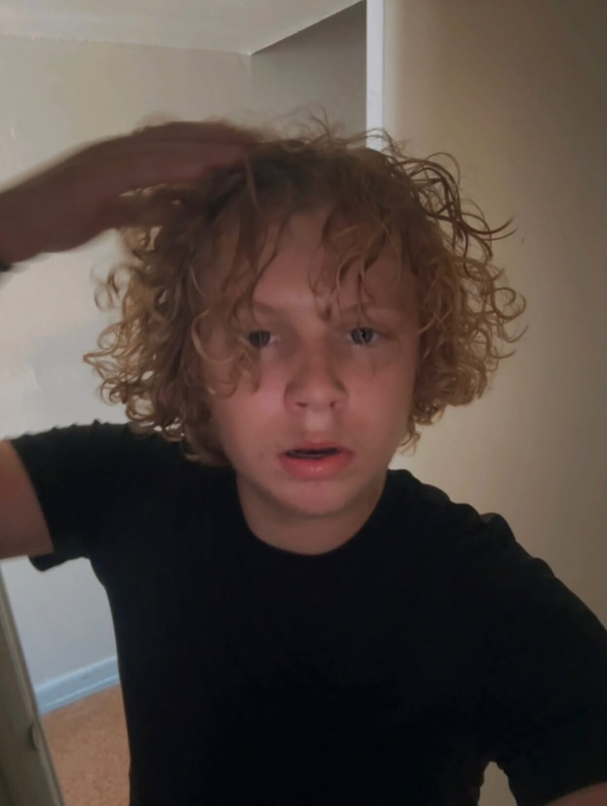 Curly-haired person in a black shirt touching their hair indoors.