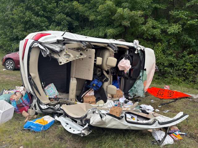 Overturned white vehicle wrecked on its side beside a wooded roadside with debris scattered around.
