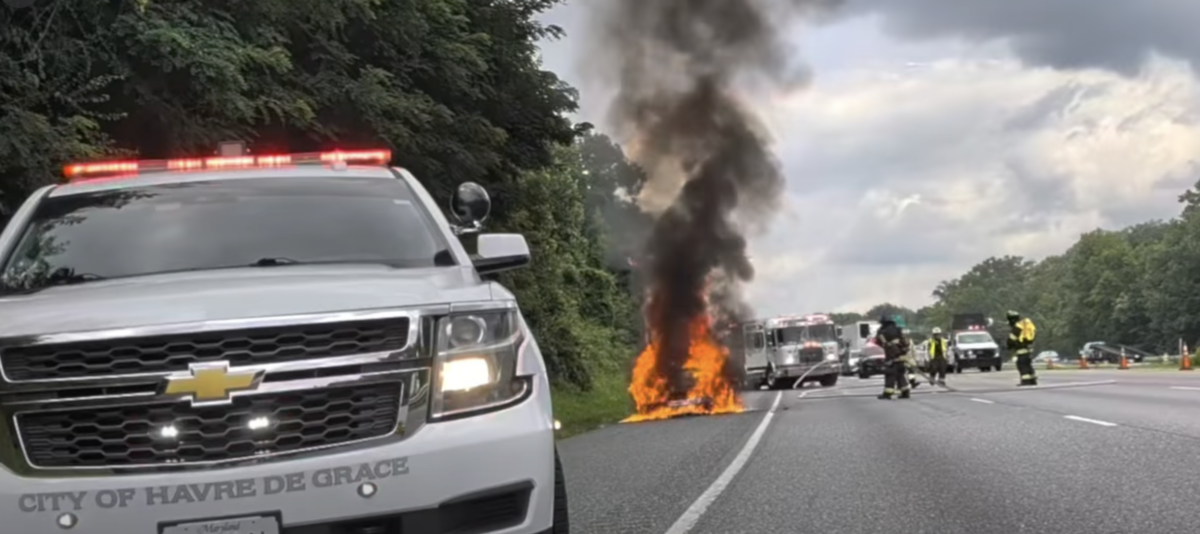 Police SUV blocks a highway while a car burns and smoke rises ahead.