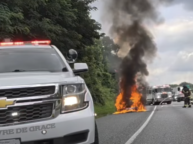 Police SUV blocks a highway while a car burns and smoke rises ahead.
