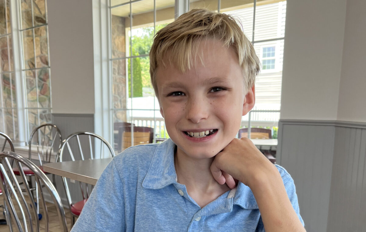 Smiling young boy in a blue shirt sitting indoors near a window.