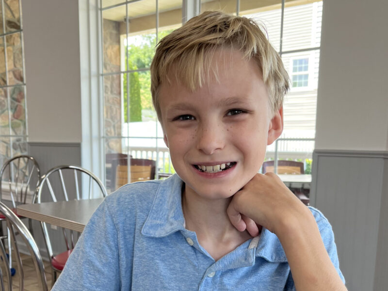 Smiling young boy in a blue shirt sitting indoors near a window.