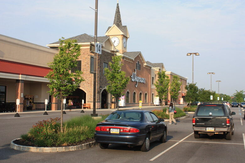 Strip mall storefront with a clock tower, parked cars, and shoppers along a sunny road.