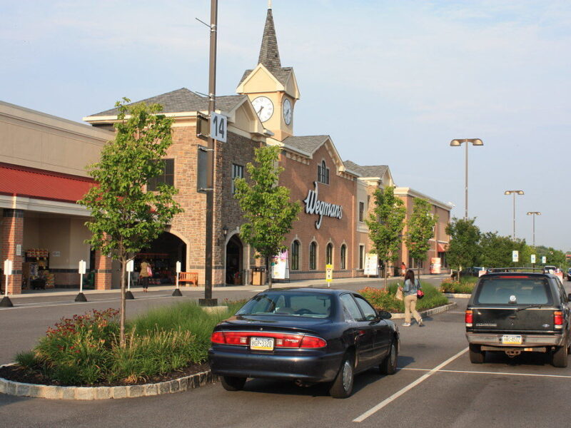 Strip mall storefront with a clock tower, parked cars, and shoppers along a sunny road.