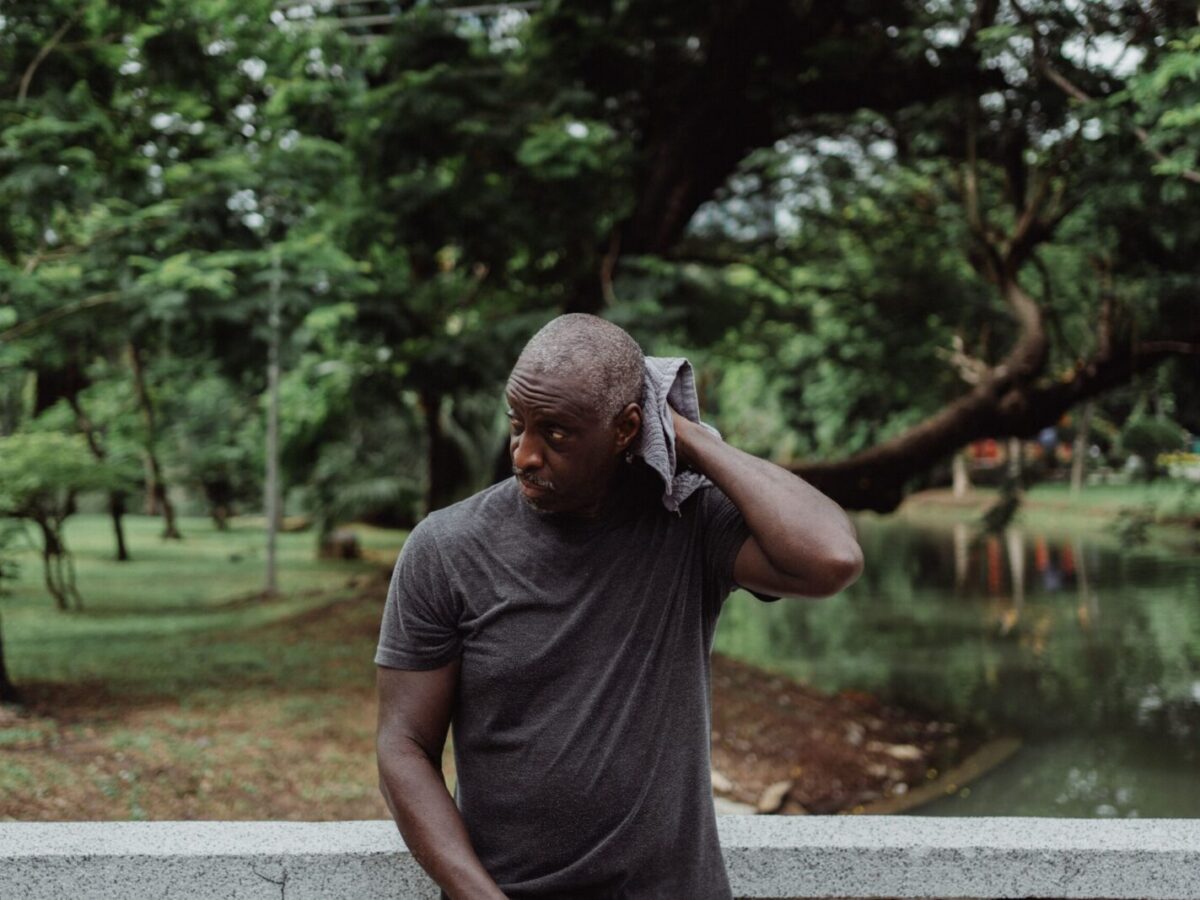 A man wipes his sweat with a towel while standing pensively outdoors in a lush park environment.