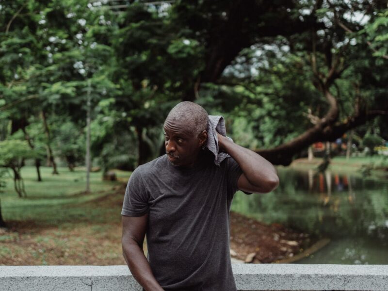 A man wipes his sweat with a towel while standing pensively outdoors in a lush park environment.