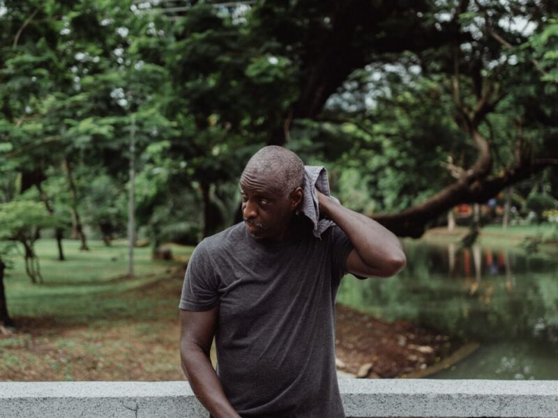 A man wipes his sweat with a towel while standing pensively outdoors in a lush park environment.
