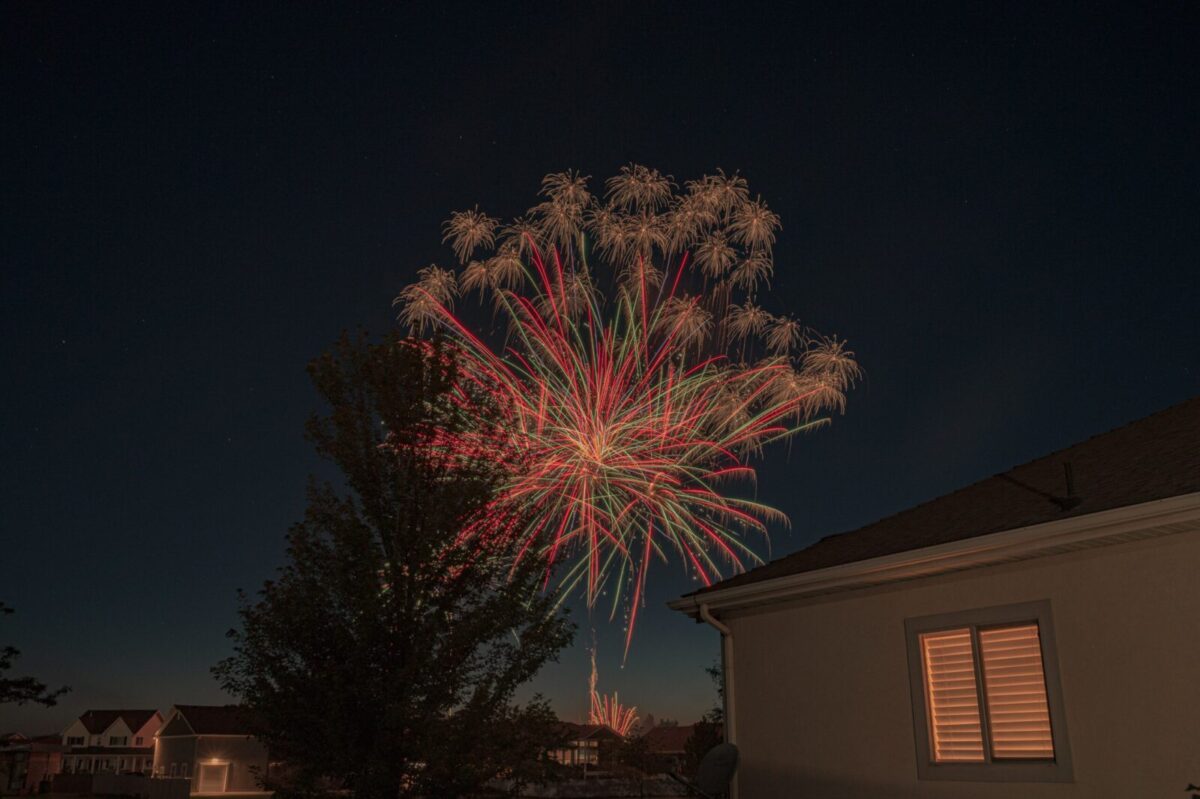 Colorful fireworks display illuminating suburban night sky with houses in the foreground.