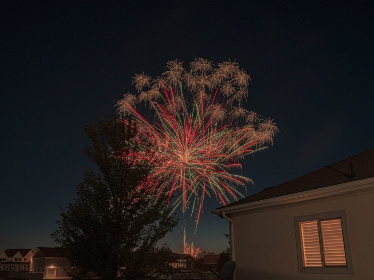 Colorful fireworks display illuminating suburban night sky with houses in the foreground.