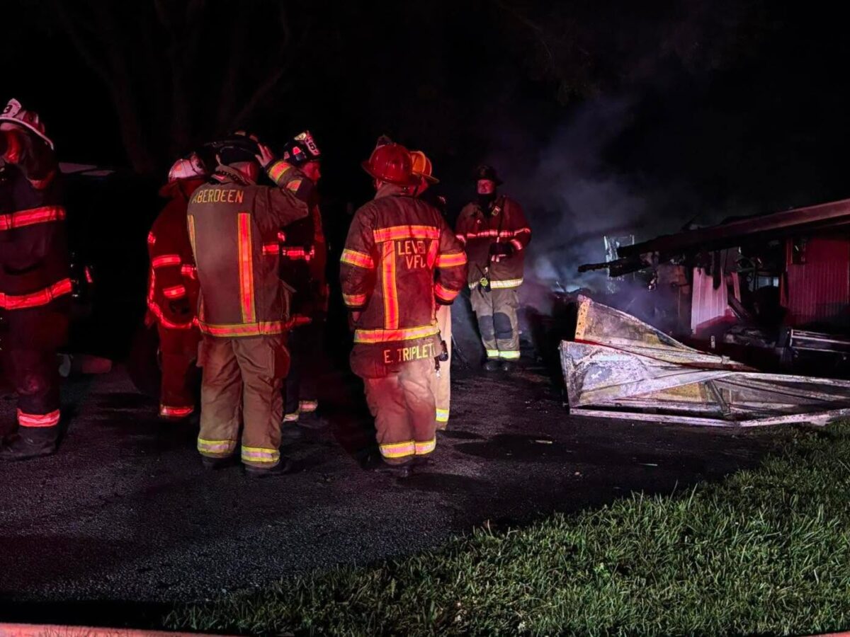Firefighters stand at a nighttime scene beside a burnt, collapsed structure lit by red emergency lights.
