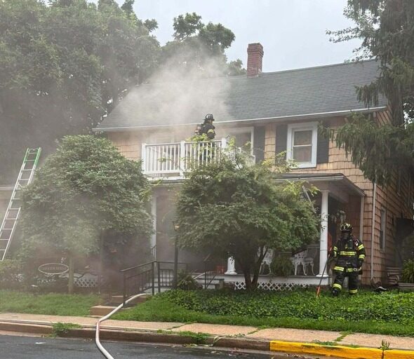 Firefighters work amid smoke at a two-story house with a ladder and hose in front.