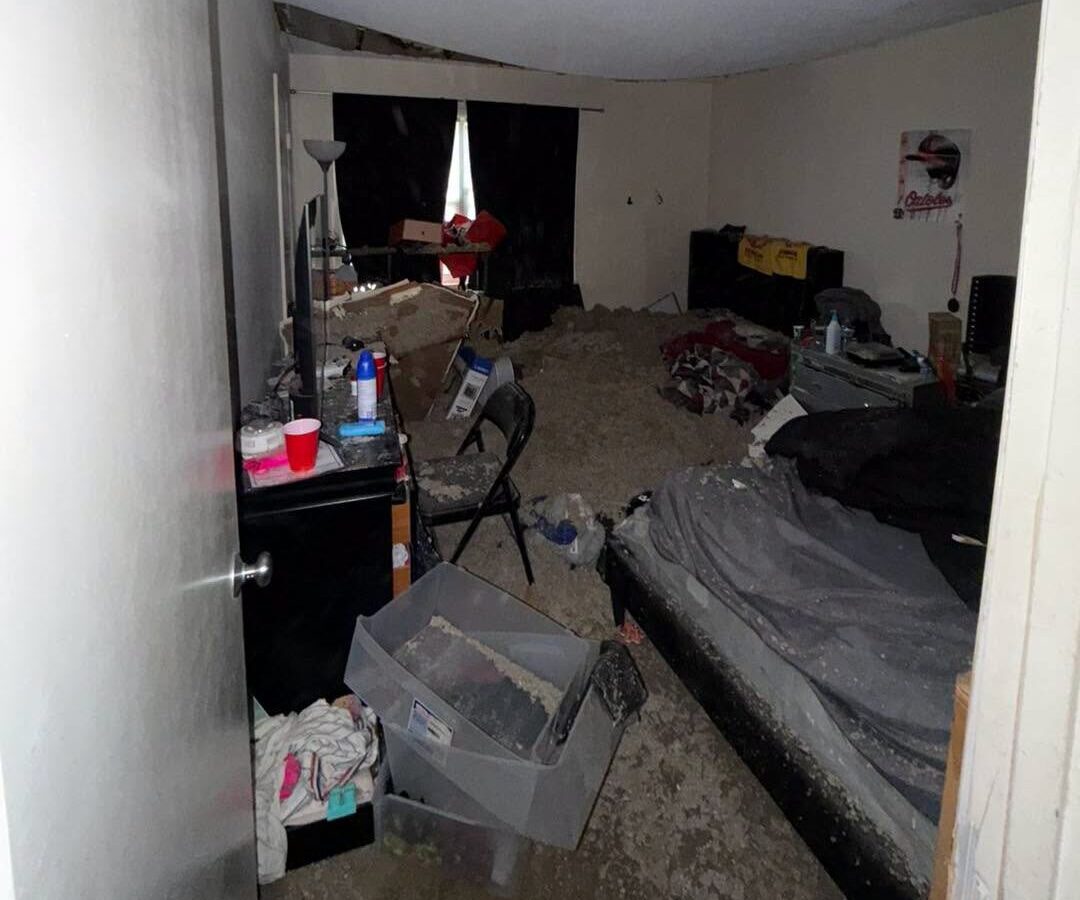 Cluttered bedroom with an unmade bed, boxes, scattered items, and a floor covered in debris.