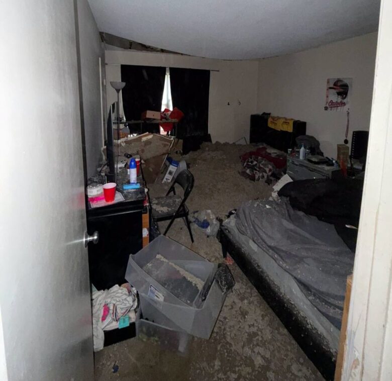 Cluttered bedroom with an unmade bed, boxes, scattered items, and a floor covered in debris.