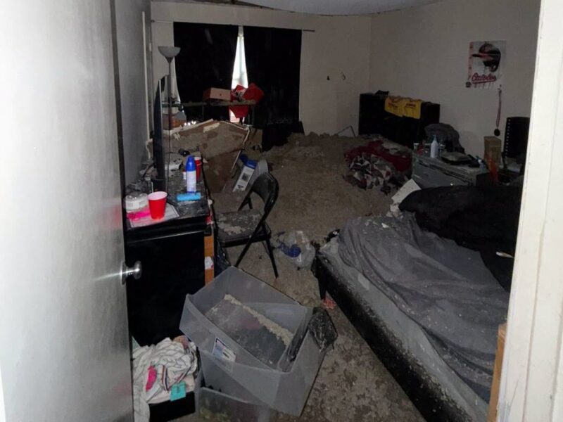 Cluttered bedroom with an unmade bed, boxes, scattered items, and a floor covered in debris.