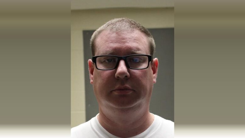 Portrait of a man wearing black glasses against a muted indoor background.
