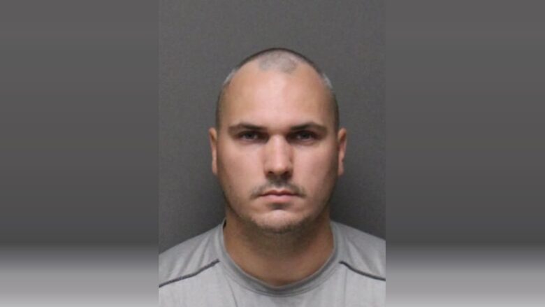 Mugshot-style portrait of a bald man in a gray shirt against a plain gray background.