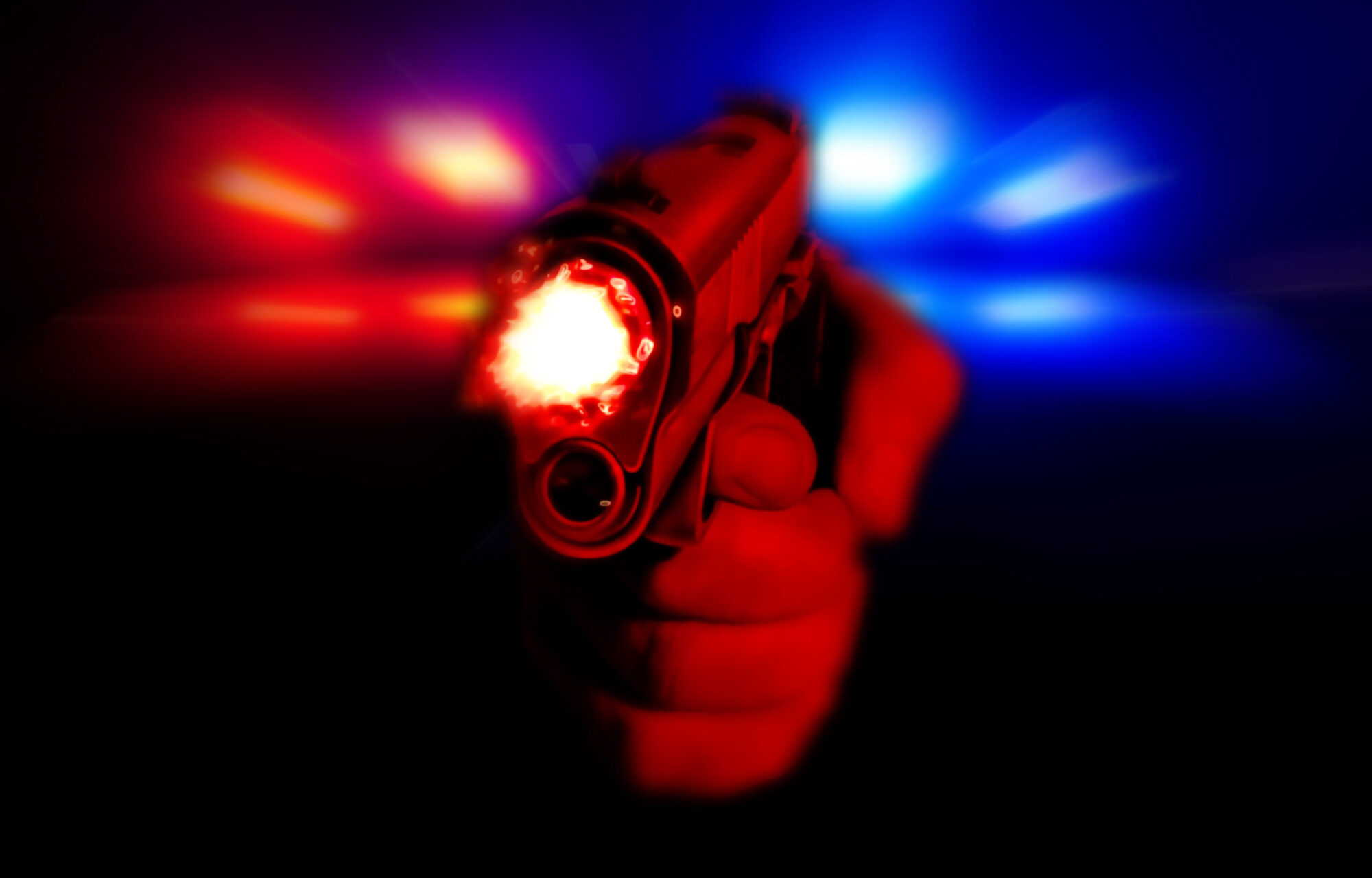 Hand holding a glowing handgun aimed at the viewer with red and blue police lights in the background.