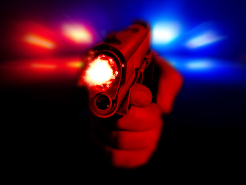 Hand holding a glowing handgun aimed at the viewer with red and blue police lights in the background.