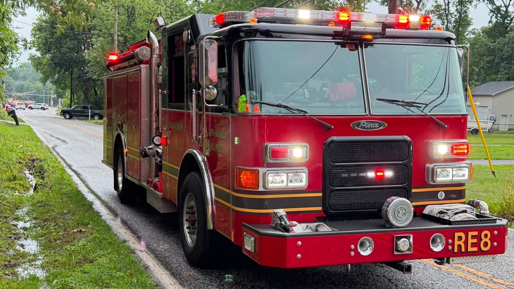 Red fire truck parked on a wet roadside with lights flashing in the rain.