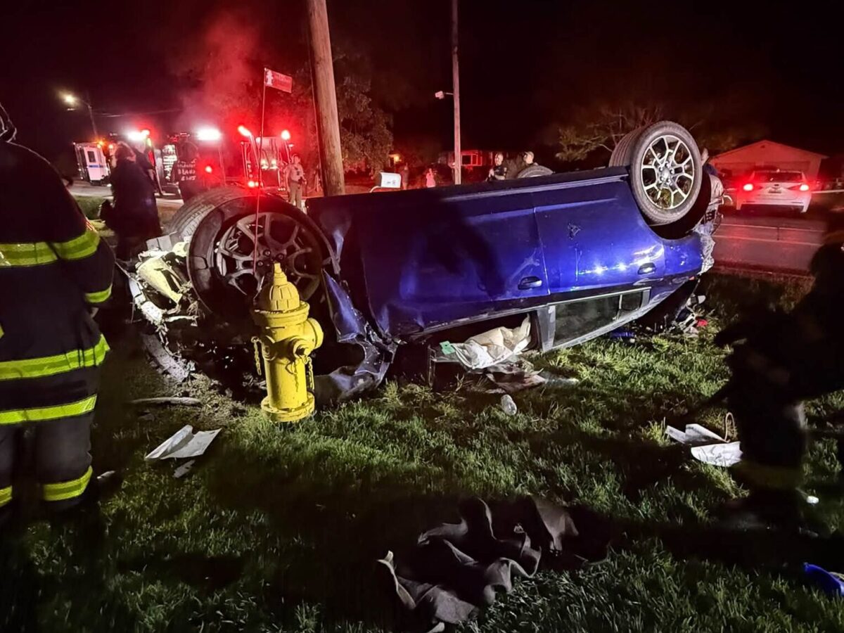 Overturned blue car beside a yellow fire hydrant at a nighttime crash scene with emergency lights.