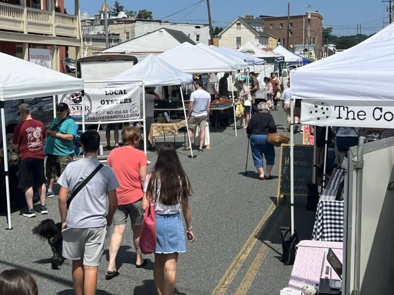 People browse white tents at a lively outdoor street market on a sunny day.