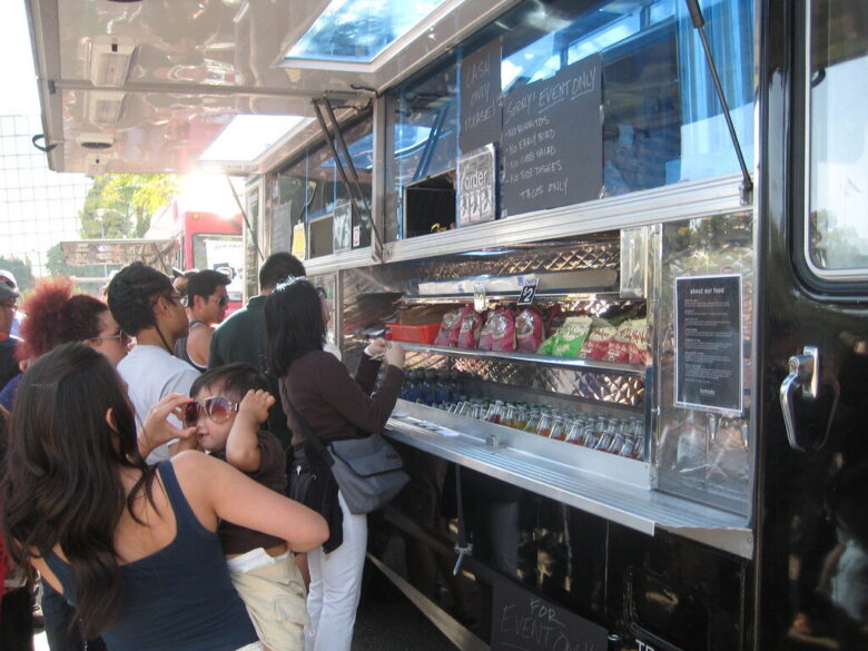 People line up at a food truck serving snacks and drinks on a sunny street.