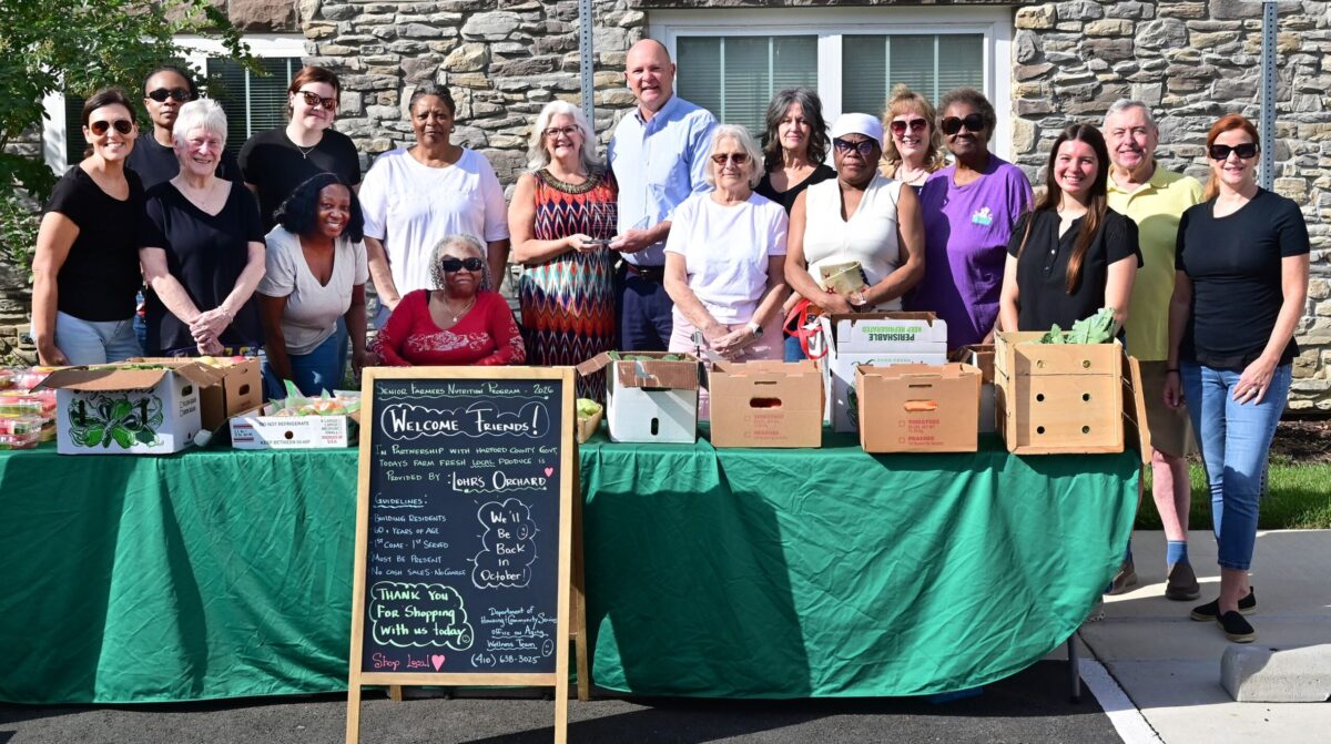 Group of people standing behind a table of boxed produce and a welcome chalkboard sign outdoors.