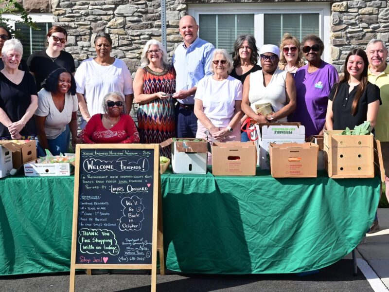 Group of people standing behind a table of boxed produce and a welcome chalkboard sign outdoors.