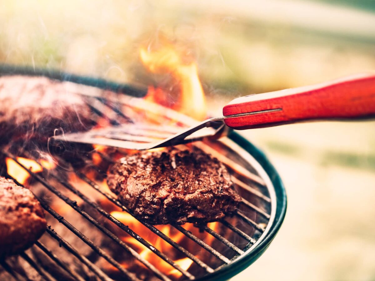 Steaks sizzling on a charcoal grill as tongs turn one over over open flames.