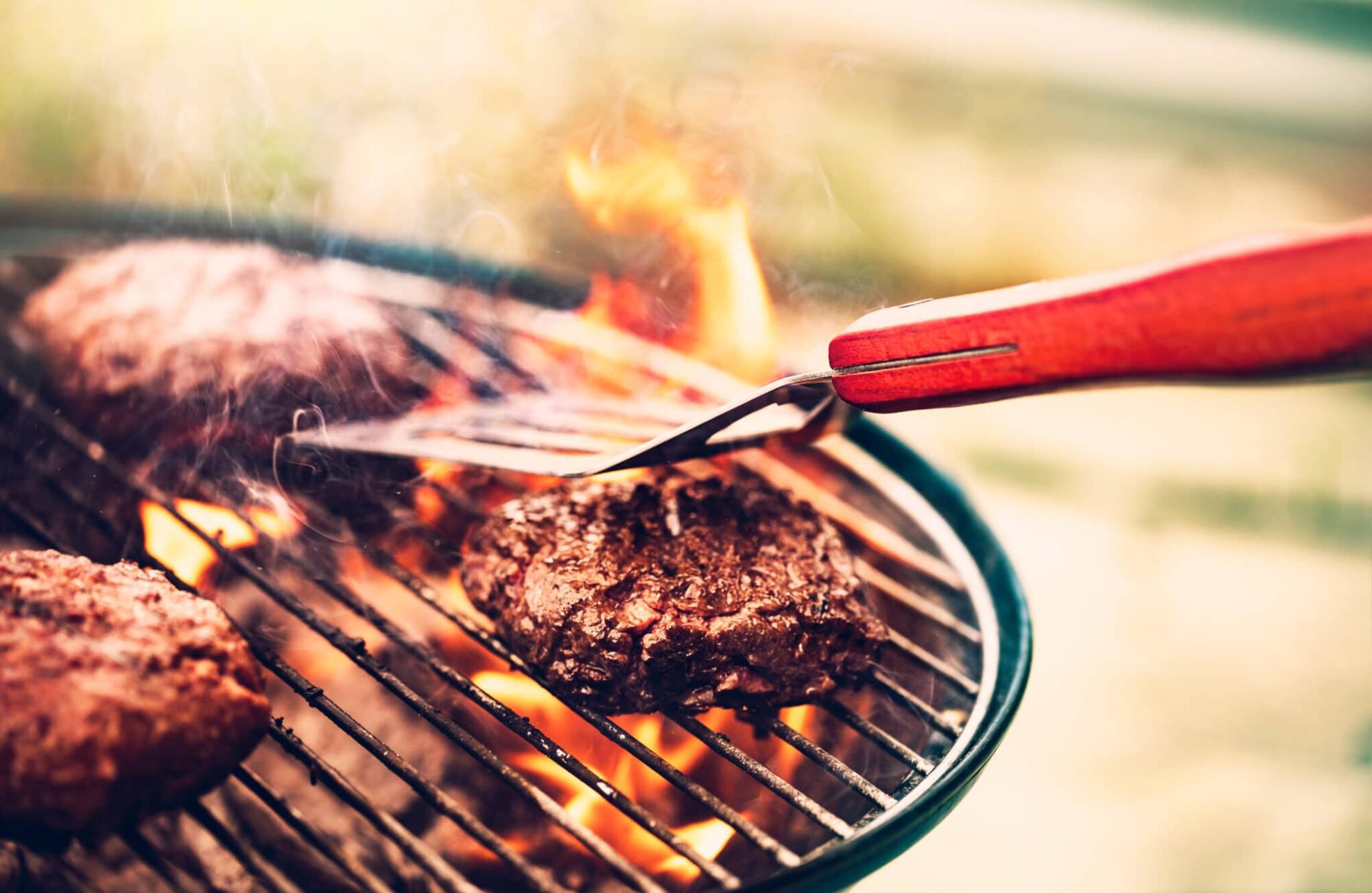 Steaks sizzling on a charcoal grill as tongs turn one over over open flames.
